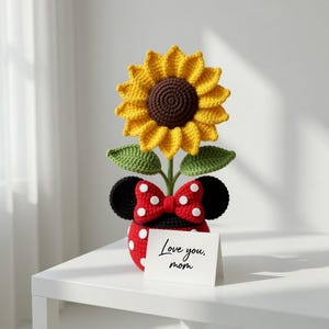May include: A crocheted sunflower in a red polka-dot pot with black ears and a red bow with white polka dots. The sunflower has yellow petals and a brown center. A small card reads "Love you, mom."