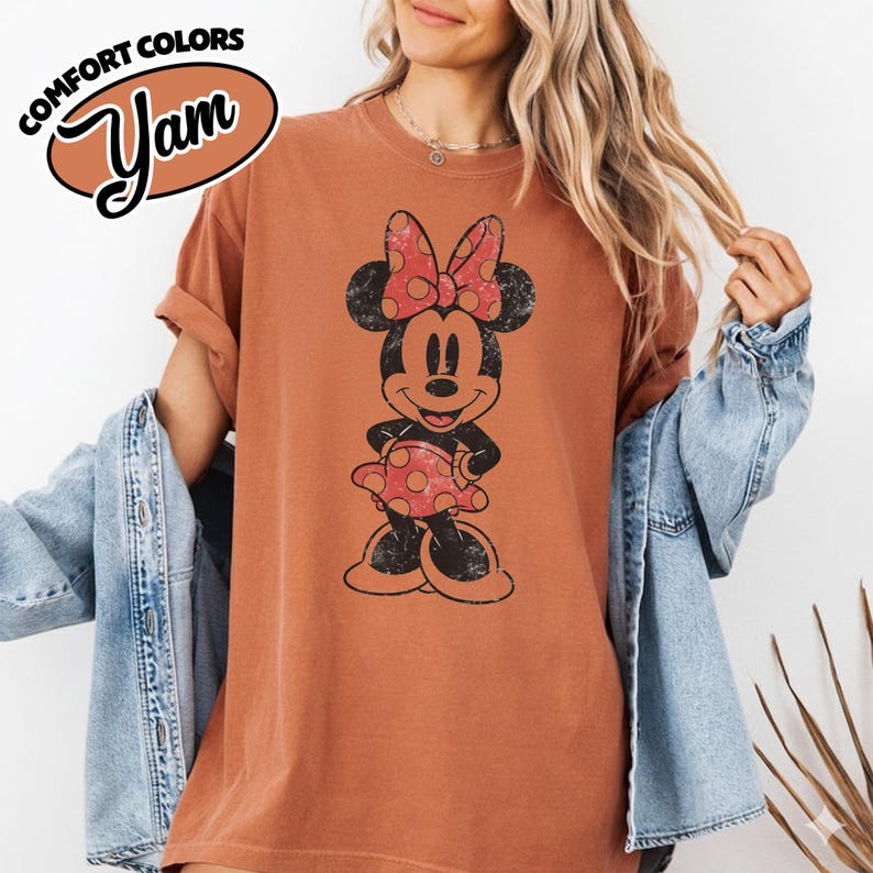 May include: A rust-colored t-shirt featuring a distressed graphic of Minnie Mouse wearing a polka-dot dress and bow. The shirt has the text "COMFORT COLORS" and "Yam" in a brown oval. The shirt has rolled-up sleeves.