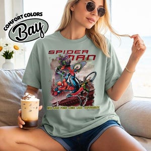 May include: A light green t-shirt featuring a Spider-Man graphic. The graphic includes the words "Spider-Man" in red and the phrase "With great power comes great responsibility." The shirt also has the words "Comfort Colors Bay" in a white oval.