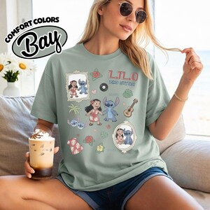 Puede incluir: Una camiseta verde salvia con un dise&ntilde;o gr&aacute;fico con las palabras "Lilo and Stitch" y varias ilustraciones de los personajes. La camiseta tambi&eacute;n tiene las palabras "Comfort Colors Bay" en un dise&ntilde;o circular.