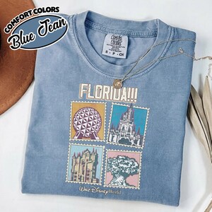 Może przedstawiać: Niebieska koszulka Comfort Colors z grafiką czterech obraz&oacute;w z Walt Disney World. Koszulka ma napis "FLORIDA!!!" wydrukowany nad obrazami. Koszulka jest złożona i ma na wierzchu naszyjnik.
