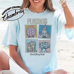 Może przedstawiać: Jasnoniebieska koszulka z grafiką przedstawiającą cztery obrazy w stylu znaczk&oacute;w pocztowych z zabytkami Florydy, w tym kulę, zamek, wieżę i drzewo. Słowo "FLORIDA!!!" jest wydrukowane nad obrazami, a "Walt Disney World" poniżej.