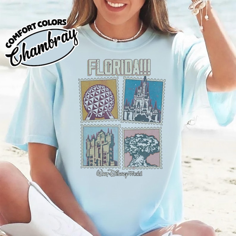 Może przedstawiać: Jasnoniebieska koszulka z grafiką przedstawiającą cztery obrazy w stylu znaczk&oacute;w pocztowych z zabytkami Florydy, w tym kulę, zamek, wieżę i drzewo. Słowo "FLORIDA!!!" jest wydrukowane nad obrazami, a "Walt Disney World" poniżej.