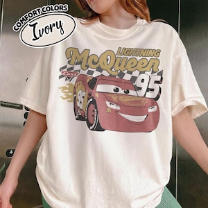 May include: Ivory-colored t-shirt featuring a graphic of Lightning McQueen from the Cars movie. The design includes the text "Lightning McQueen 95" and a checkered flag. The shirt is labeled "Comfort Colors" and is a short-sleeved tee.