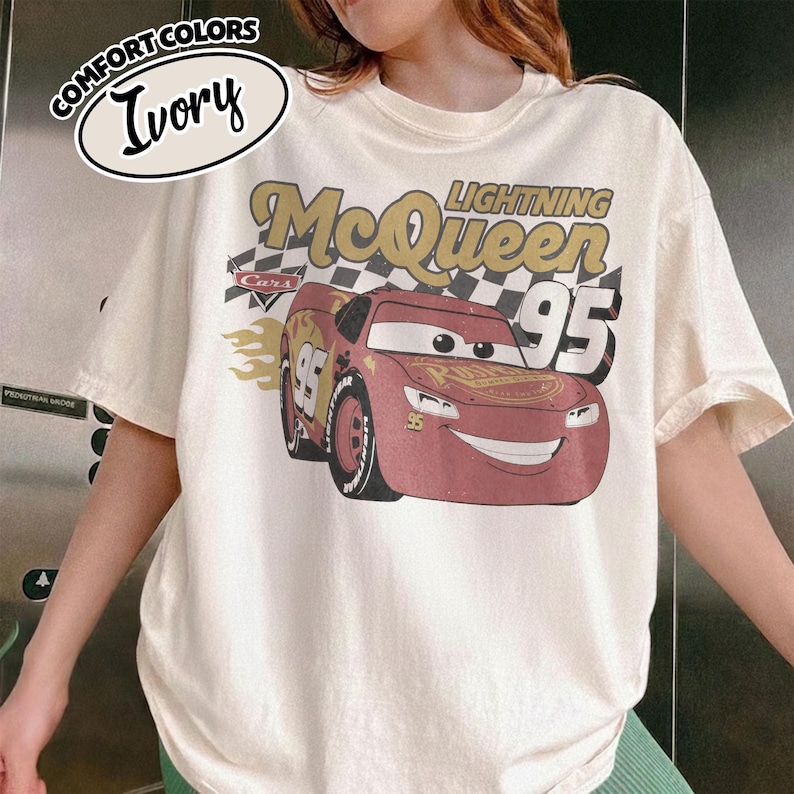 May include: Ivory-colored t-shirt featuring a graphic of Lightning McQueen from the Cars movie. The design includes the text "Lightning McQueen 95" and a checkered flag. The shirt is labeled "Comfort Colors" and is a short-sleeved tee.