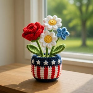 May include: A crocheted floral arrangement featuring a red rose, white daisies with yellow centers, and a blue flower. The flowers are in a blue pot with white stars and red and white stripes, resembling a patriotic theme.