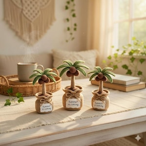 May include: Three crochet palm tree decorations with green fronds and brown coconuts in burlap pots. Each pot has a tag that reads "Summer is coming". The trees are on a white table.