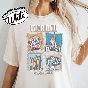 Może przedstawiać: Biała koszulka Comfort Colors z grafiką z napisem "FLORIDA!!!" i czterema obrazami w stylu znaczka pocztowego z zabytkami Florydy, w tym geodezyjną kulą, zamkiem i drzewem. U dołu napis "Walt Disney World".