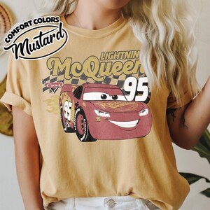 May include: Mustard-colored t-shirt featuring a graphic of Lightning McQueen from the Cars movie. The design includes the number 95, the name "Lightning McQueen," and the word "Cars." The shirt has rolled-up sleeves and a vintage aesthetic.
