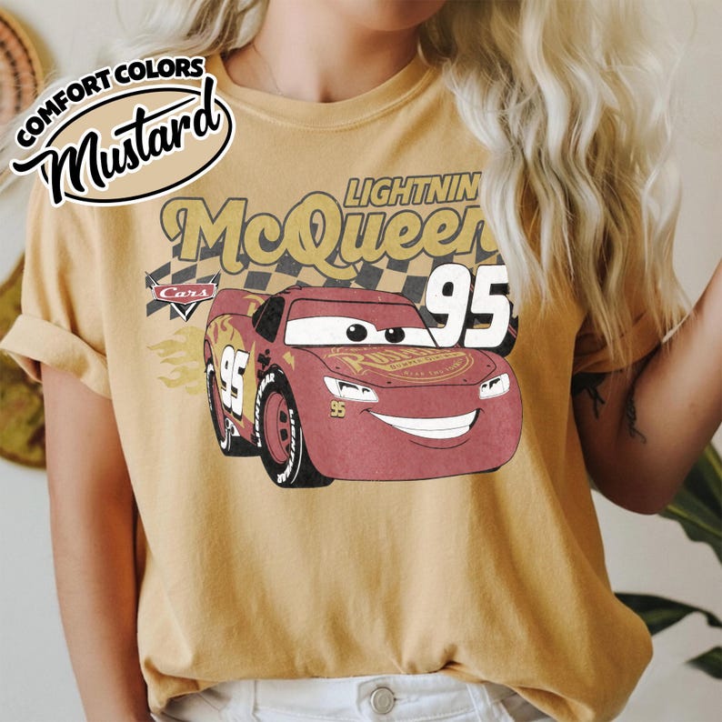 May include: Mustard-colored t-shirt featuring a graphic of Lightning McQueen from the Cars movie. The design includes the number 95, the name "Lightning McQueen," and the word "Cars." The shirt has rolled-up sleeves and a vintage aesthetic.