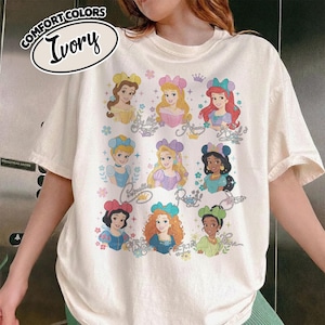 May include: Ivory-colored t-shirt featuring a graphic of various cartoon princesses. The shirt has a crew neck and short sleeves. The text "COMFORT COLORS" and "Ivory" are printed on the top left of the shirt.
