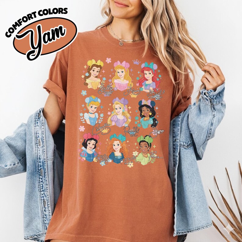 May include: A rust-colored t-shirt featuring illustrations of various princesses. The shirt has the words "Comfort Colors" and "Yam" printed on the top left. The princesses are depicted with floral accents and their names.