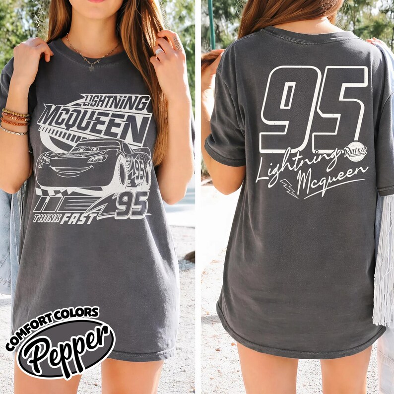 May include: Charcoal grey t-shirt with a Lightning McQueen design. The front shows the character, number 95, and "Think Fast." The back has a large white "95" and "Lightning McQueen" text. The shirt is a Comfort Colors Pepper style.