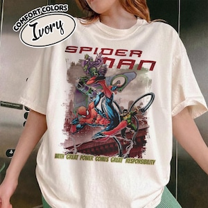 May include: Ivory-colored t-shirt featuring a Spider-Man graphic. The design includes Spider-Man in action, along with the text "SPIDER-MAN" and the phrase "WITH GREAT POWER COMES GREAT RESPONSIBILITY". The shirt has a crew neck and short sleeves.