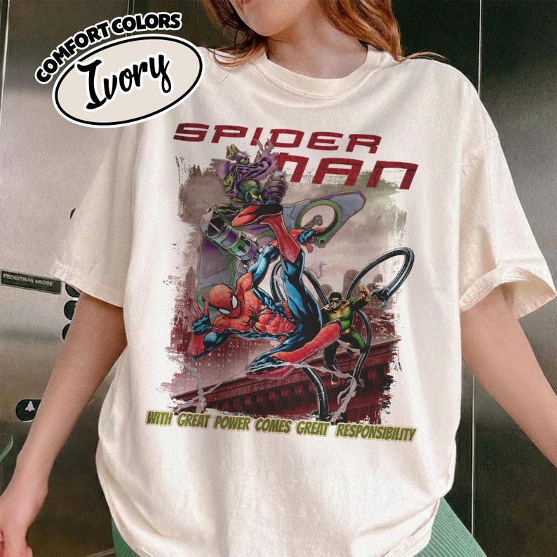 May include: Ivory-colored t-shirt featuring a Spider-Man graphic. The design includes Spider-Man in action, along with the text "SPIDER-MAN" and the phrase "WITH GREAT POWER COMES GREAT RESPONSIBILITY". The shirt has a crew neck and short sleeves.