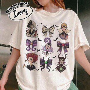 May include: Ivory-colored t-shirt with a graphic print featuring cartoon illustrations of villainous characters and bows. The shirt has the words "Comfort Colors" and "Ivory" printed on the top left corner.