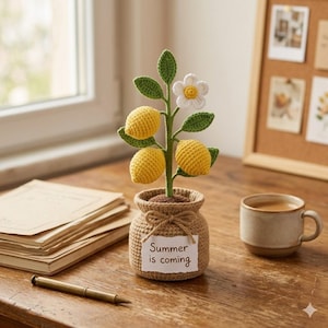 May include: A crocheted lemon tree with yellow lemons, green leaves, and a white flower in a small woven pot. The pot has a tag that reads "Summer is coming". The tree sits on a wooden surface next to a cup of coffee and a stack of books.