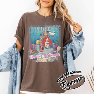 Puede incluir: Camiseta color espresso con un gr&aacute;fico colorido con el texto "I Wanna Be Where The People Aren't". El gr&aacute;fico incluye un castillo, una sirena y otras criaturas marinas. Parte de la colecci&oacute;n Comfort Colors.