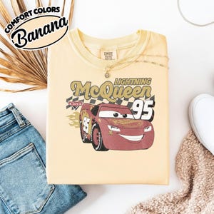 May include: A yellow Comfort Colors t-shirt with a graphic of Lightning McQueen from the Cars movie. The design features the text "Lightning McQueen 95" and a checkered flag background. The shirt is folded on a white surface.