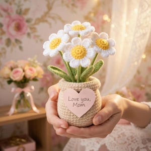 May include: A handmade crochet flower arrangement featuring white daisies with yellow centers and green stems, nestled in a beige crocheted pot. A pink heart-shaped tag reads "Love you, Mom".