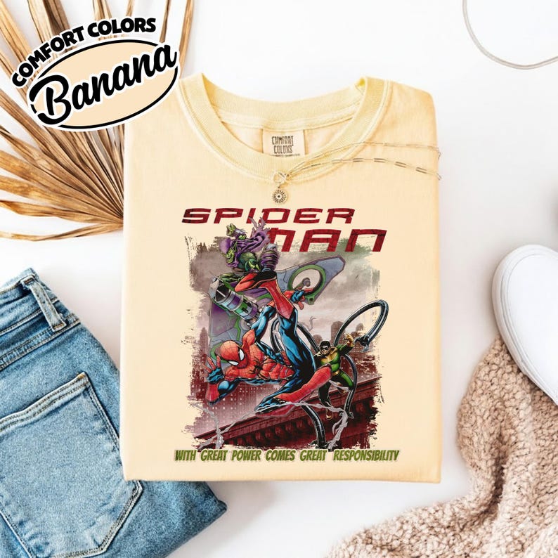 May include: A light yellow t-shirt featuring a Spider-Man graphic. The graphic includes Spider-Man, the Green Goblin, and other characters. The text "SPIDER-MAN" is above the graphic, and "WITH GREAT POWER COMES GREAT RESPONSIBILITY" is below.