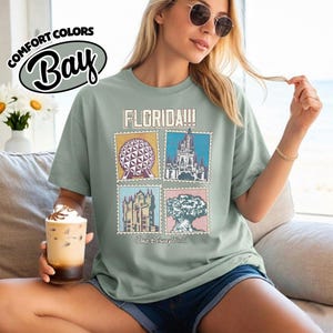 Może przedstawiać: Szałwiowa zielona koszulka z grafiką Florydy przedstawiającą cztery obrazy w stylu znaczka pocztowego z zabytkami Disney World. Koszulka ma napis "FLORIDA!!!" i "Walt Disney World". Koszulka jest wykonana z materiału Comfort Colors.