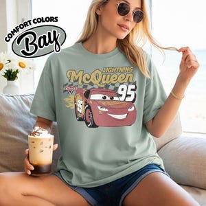 May include: Sage green t-shirt featuring a graphic of Lightning McQueen from the Cars movie. The design includes the text "Lightning McQueen 95" in a vintage style. The shirt is from Comfort Colors Bay.
