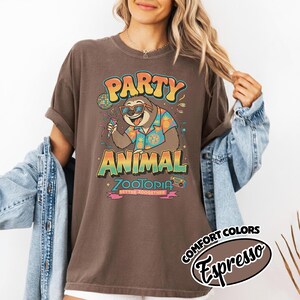May include: Brown t-shirt with a graphic of a sloth wearing sunglasses and a Hawaiian shirt, holding a microphone. The text reads "PARTY ANIMAL ZOOTOPIA BETTER ZOOOGETHER". The shirt is labeled "Comfort Colors Espresso".
