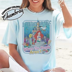 Puede incluir: Camiseta azul claro con un gr&aacute;fico de Ariel de La Sirenita, un castillo y el texto "I Wanna Be Where The People Aren't". La camiseta tiene las palabras "Comfort Colors Chambray" en la esquina superior izquierda.
