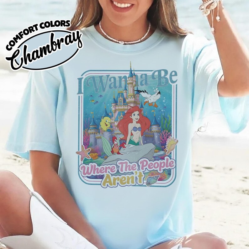 Puede incluir: Camiseta azul claro con un gr&aacute;fico de Ariel de La Sirenita, un castillo y el texto "I Wanna Be Where The People Aren't". La camiseta tiene las palabras "Comfort Colors Chambray" en la esquina superior izquierda.