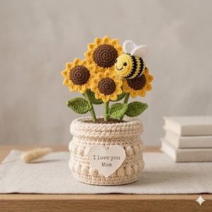 May include: A crocheted sunflower arrangement in a beige pot, featuring yellow and brown sunflowers, green leaves, and a bee. The pot has a heart-shaped tag that reads "I love you Mom."