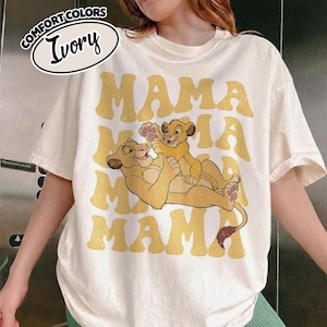 May include: Ivory-colored t-shirt with the word "MAMA" in yellow retro font. The shirt features a cartoon illustration of a lioness and cub. The top left corner has a label that says "Comfort Colors" and "Ivory".