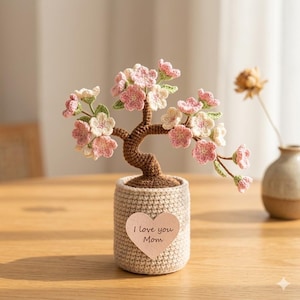 May include: A crocheted bonsai tree with pink and white flowers in a beige pot. A heart-shaped tag on the pot reads "I love you Mom". The tree has a brown trunk and green leaves, creating a charming, handmade decorative item.