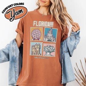 Może przedstawiać: Rdzawa koszulka z grafiką przedstawiającą cztery obrazy w stylu znaczk&oacute;w pocztowych z zabytkami Florydy, w tym geometryczną kulę, zamek, budynek i drzewo. Tekst "FLORIDA!!!" znajduje się nad obrazami, a "Walt Disney World!" poniżej.