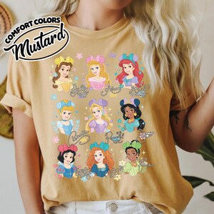 May include: Mustard-colored t-shirt featuring illustrations of various princesses. Each princess is depicted with unique hairstyles and outfits, with their names written below. The shirt has the words "Comfort Colors" and "Mustard" printed on the top left.