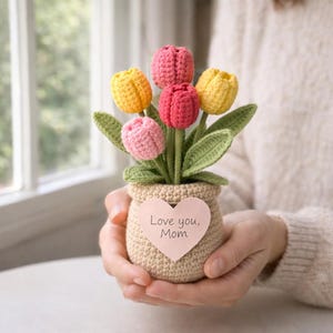 May include: A crocheted bouquet of tulips in shades of pink and yellow, with green leaves, sits in a small beige pot. A heart-shaped tag on the pot reads "Love you, Mom."