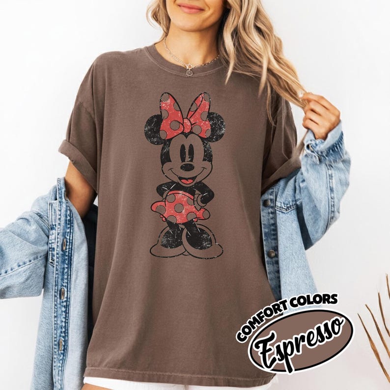 May include: Espresso-colored t-shirt featuring a distressed Minnie Mouse graphic with a red polka-dot bow and dress. The shirt has rolled-up sleeves and a Comfort Colors label.