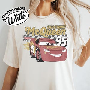 May include: White Comfort Colors t-shirt featuring a graphic of Lightning McQueen from the Cars movie. The design includes the text "Lightning McQueen 95" and a checkered flag background. The shirt has a crew neck and short sleeves.