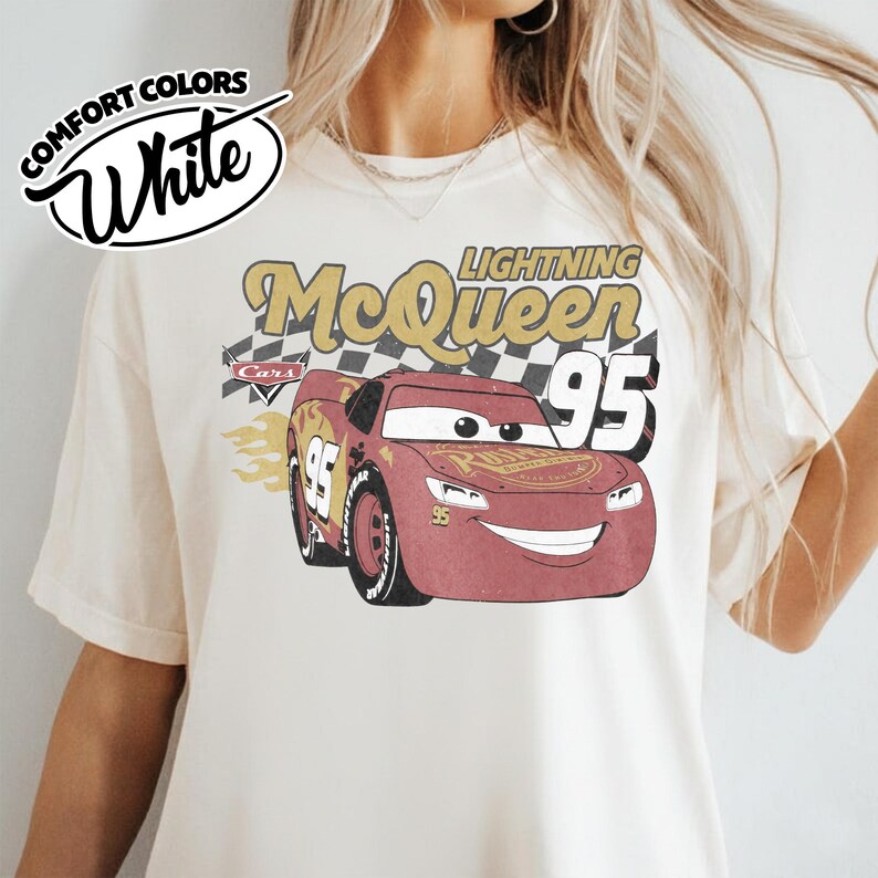 May include: White Comfort Colors t-shirt featuring a graphic of Lightning McQueen from the Cars movie. The design includes the text "Lightning McQueen 95" and a checkered flag background. The shirt has a crew neck and short sleeves.