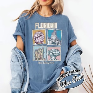 Może przedstawiać: Niebieska koszulka Comfort Colors z napisem "FLORIDA!!!" nad czterema obrazami w stylu znaczka pocztowego z zabytkami Walt Disney World. Koszulka ma podwinięte rękawy, a na okrągłej grafice widnieje napis "COMFORT COLORS Blue Jean".