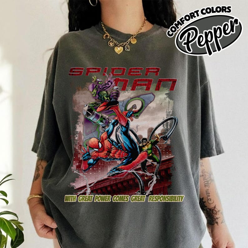 May include: Dark gray t-shirt featuring a Spider-Man graphic. The design includes Spider-Man battling the Green Goblin, with the text "SPIDER-MAN" and "WITH GREAT POWER COMES GREAT RESPONSIBILITY". The shirt has a relaxed fit and the words "COMFORT COLORS Pepper" are printed on the top right.