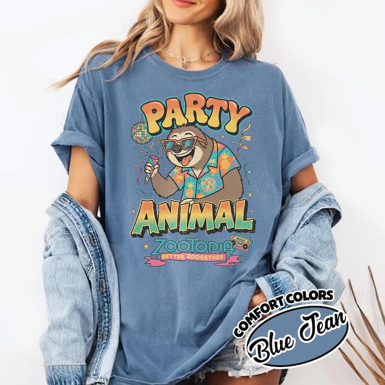 May include: Blue t-shirt with a graphic of a sloth wearing sunglasses and a Hawaiian shirt, holding a microphone. The text reads "PARTY ANIMAL Zootopia" in colorful, retro-style lettering. The shirt is labeled "Comfort Colors Blue Jean".