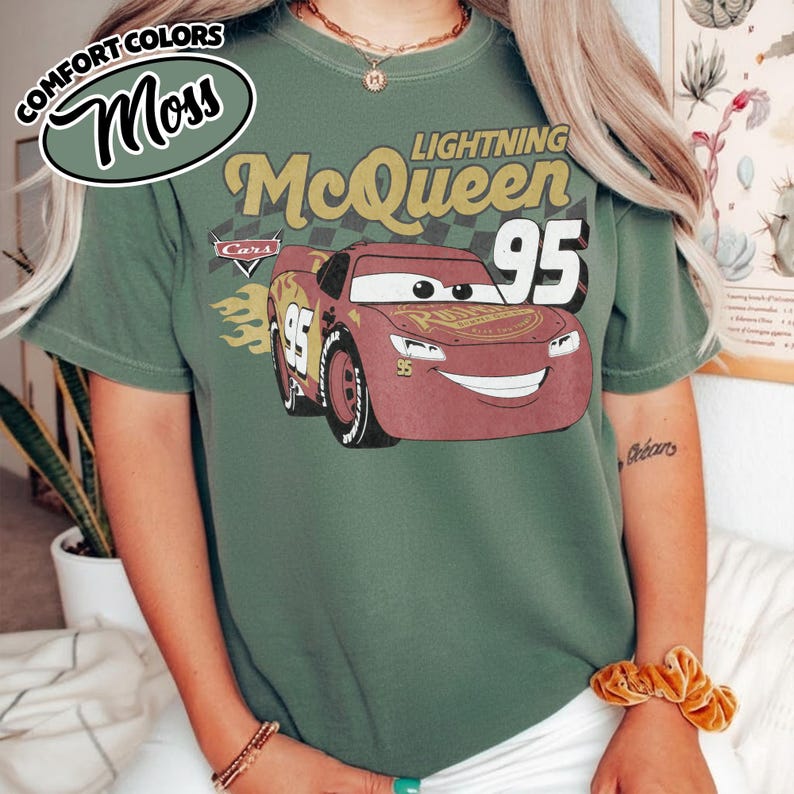 May include: Moss green t-shirt featuring Lightning McQueen from the Cars movie. The design includes the number 95, flames, and the Cars logo. The shirt has a vintage look with the words "Comfort Colors" and "Moss" in a circular design.