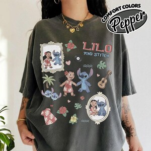 Puede incluir: Camiseta gris oscuro con el texto "Lilo and Stitch" e ilustraciones de dibujos animados de Lilo y Stitch. La camiseta incluye im&aacute;genes de una palmera, flores, una guitarra y un disco. Las palabras "Comfort Colors" y "Pepper" tambi&eacute;n est&aacute;n impresas en la camiseta.