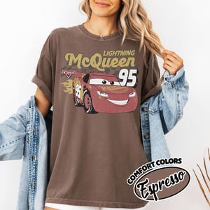 May include: Brown t-shirt featuring a graphic of Lightning McQueen from the Cars movie. The design includes the text "Lightning McQueen 95" and a stylized image of the race car. The shirt has rolled-up sleeves and a distressed vintage look.