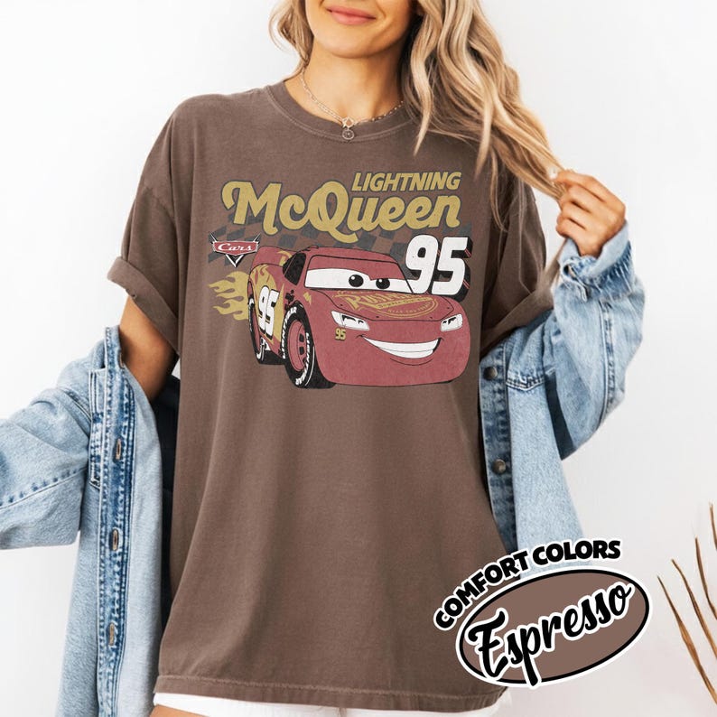 May include: Brown t-shirt featuring a graphic of Lightning McQueen from the Cars movie. The design includes the text "Lightning McQueen 95" and a stylized image of the race car. The shirt has rolled-up sleeves and a distressed vintage look.