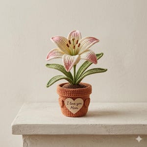 May include: A crocheted lily flower in a terracotta-colored pot. The flower has white petals with pink edges, green leaves, and a brown center. A heart-shaped tag on the pot reads "I love you Mom."