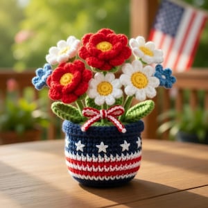 May include: A handcrafted crochet flower arrangement in a blue, red, and white pot. The flowers are red, white, and blue, with green leaves and a red and white bow. An American flag is visible in the background.