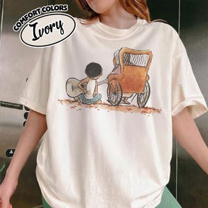 May include: Ivory-colored t-shirt featuring a whimsical illustration of a person playing a guitar for another person in a vintage carriage. The shirt has the words "Comfort Colors" and "Ivory" in a circular design at the top.