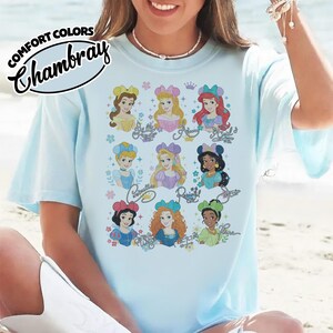 May include: Light blue t-shirt with a graphic of nine Disney princesses. The princesses are depicted with their names in cursive script. The shirt has the words "COMFORT COLORS" and "Chambray" printed on the upper left side.